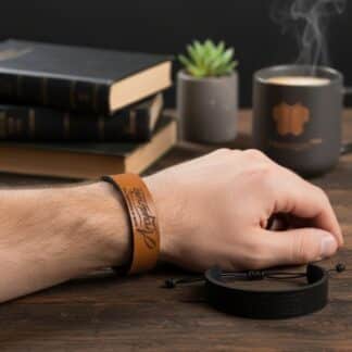 Bransoletka męska skórzana z grawerem "Arcydzieło" w kolorze brązowym na nadgarstku. W tle kubek kawy i książki.