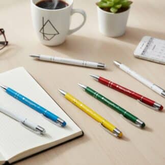 Eleganckie, metalowe długopisy w różnych kolorach (czerwony, zielony, żółty, niebieski, biały) leżące na drewnianym biurku obok otwartego notesu, okularów, filiżanki kawy i telefonu. Na długopisach widoczne jest logo Heksagon PRO i Klub 555. To ujęcie lifestylowe, idealne na grafikę produktową w sklepie internetowym.